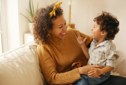 Indoor shot of happy young Hispanic woman with brown wavy hair relaxing at home embracing her adorable toddler son. Cheerful mother bonding with infant son, sitting on sofa in living room, laughing