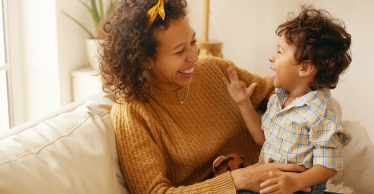 Indoor shot of happy young Hispanic woman with brown wavy hair relaxing at home embracing her adorable toddler son. Cheerful mother bonding with infant son, sitting on sofa in living room, laughing