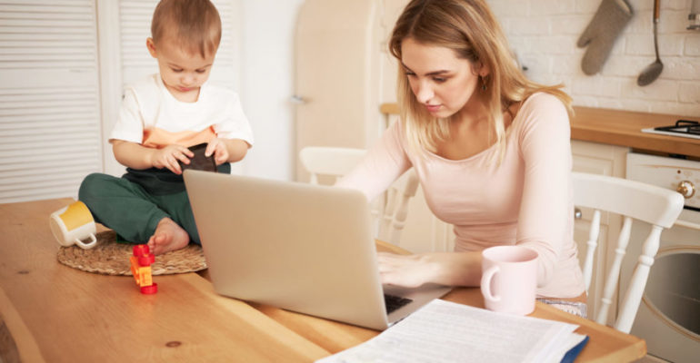Worried upset young blonde female sitting at kitchen table with papers and portable computer feeling stressed because she has to make report and take care of her baby son while he is staying at home